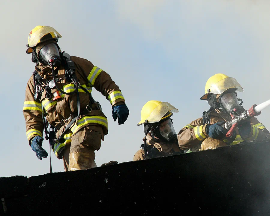 Three firefighters in yellow helmets working on a smoking roof, one aiming a hose stream.